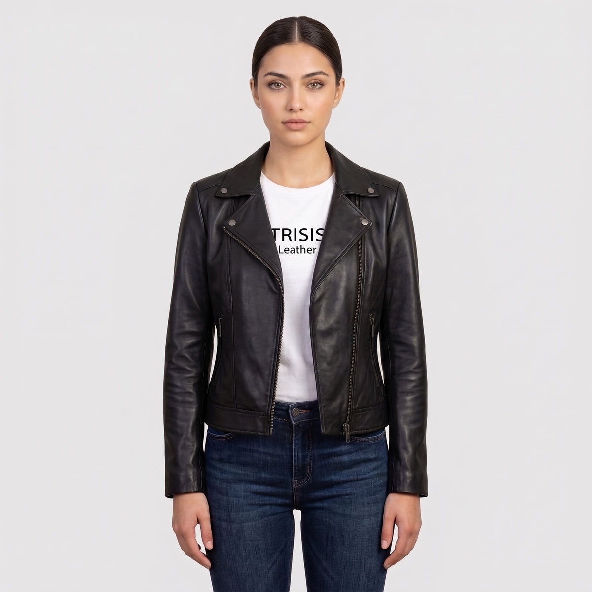 A woman modeling a black leather moto jacket over a white graphic t-shirt and dark blue jeans, shot against a solid light gray background.